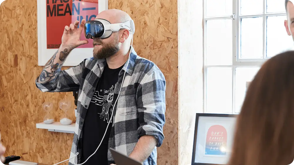 A bearded man wearing a plaid shirt and VR headset stands indoors near a window, holding the headset with one hand. Posters and framed art decorate the wooden wall behind him.