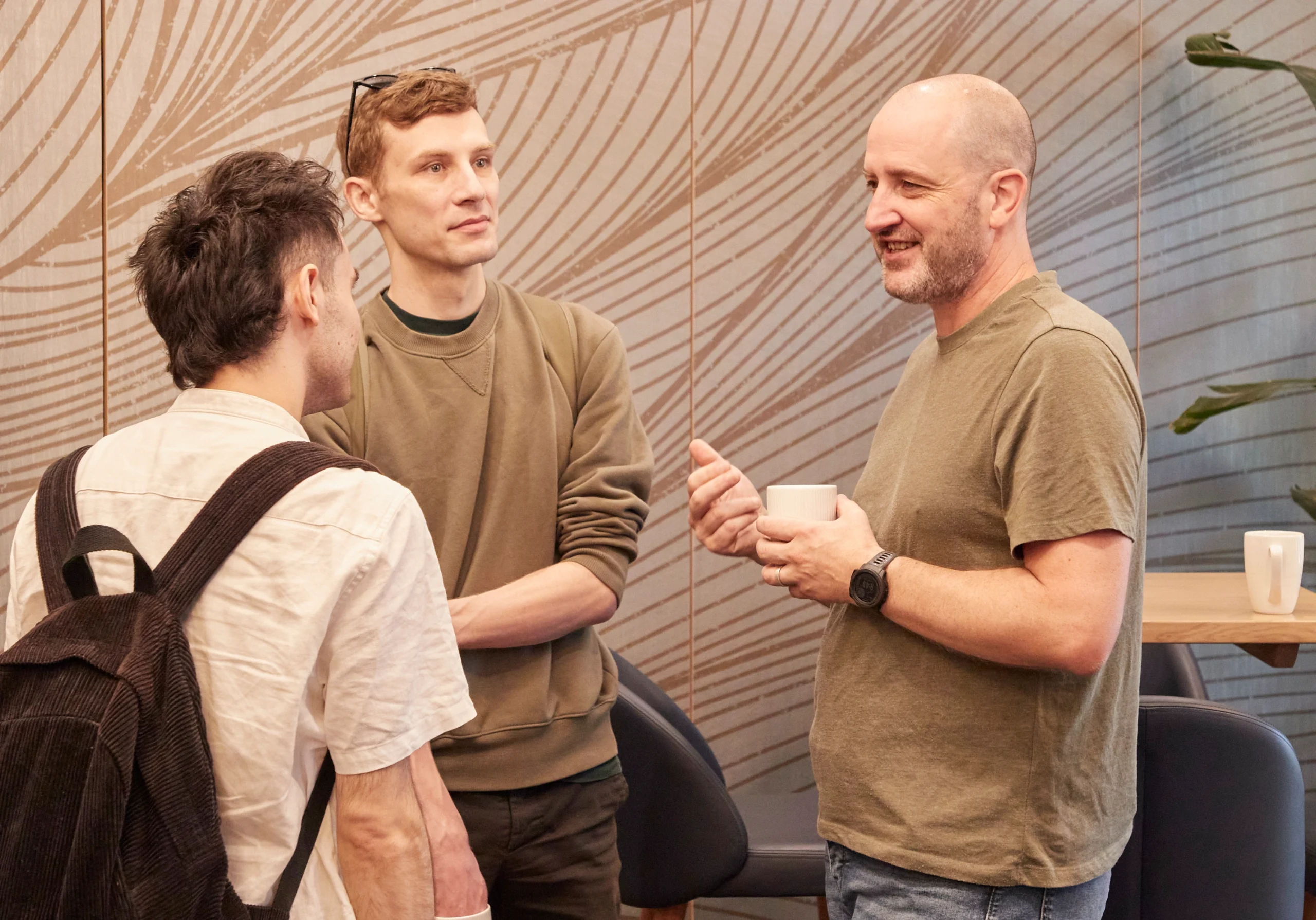 Three men are standing and talking in a casual indoor setting. One holds a white mug, another has his arms folded, and the third wears a rucksack. They appear to be engaged in a friendly conversation.