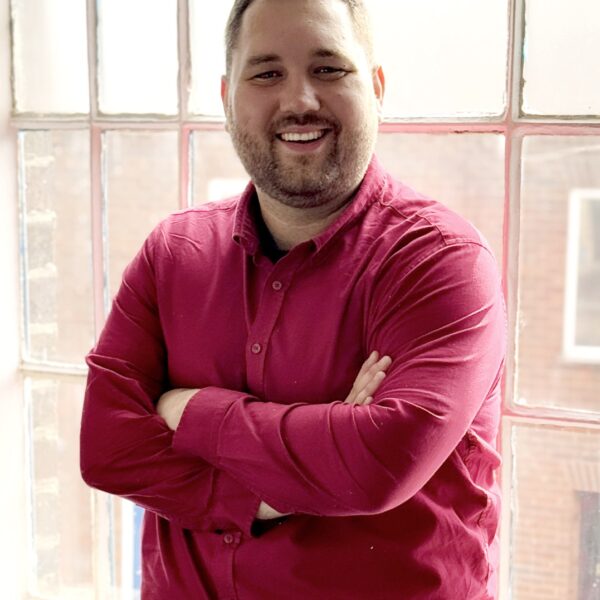 A man with short hair and a beard, wearing a bright pink button-up shirt, stands smiling with arms crossed in front of a large window with frosted glass, letting in natural light.