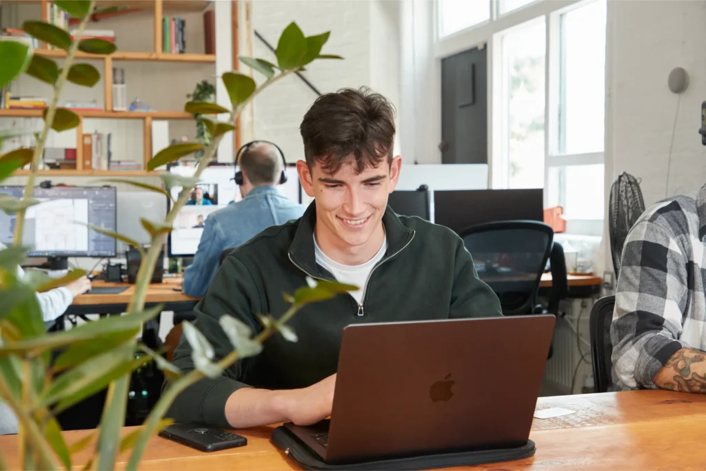 A young man smiles whilst working on a laptop at a wooden desk in a bright, modern office with colleagues in the background and a leafy plant in the foreground.