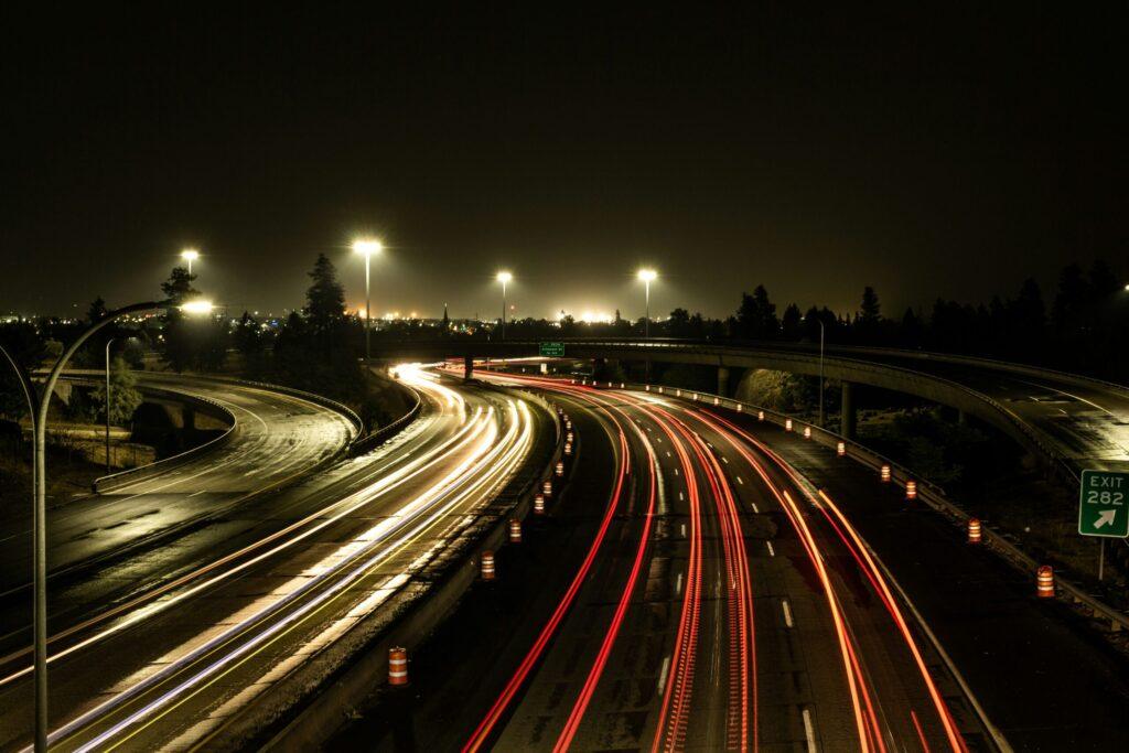 A night-time view of a motorway interchange with light trails from moving vehicles, streetlights illuminating the scene, and an exit sign for 282 visible on the right. Trees and distant city lights are in the background.