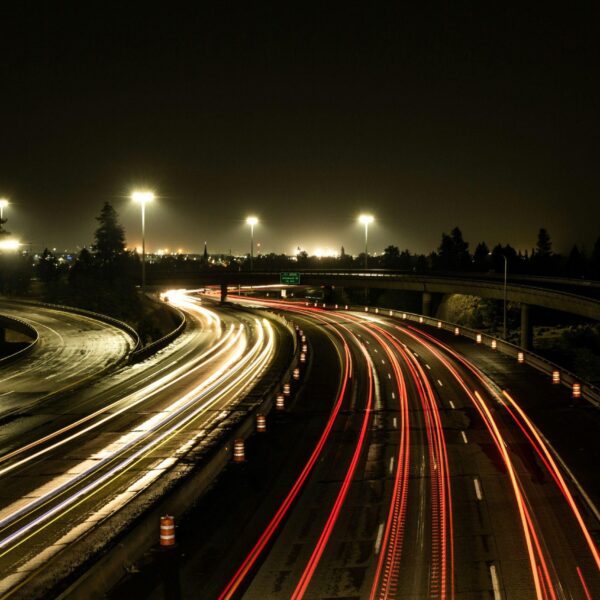 A night-time view of a motorway interchange with light trails from moving vehicles, streetlights illuminating the scene, and an exit sign for 282 visible on the right. Trees and distant city lights are in the background.