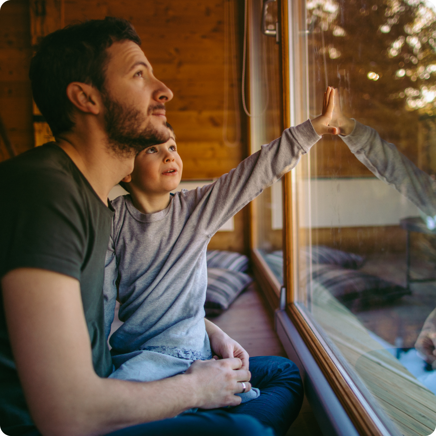 A man sits with a young boy by a large window. The boy reaches out to touch the glass, looking outside with curiosity, while the man gazes in the same direction. Warm wooden walls and natural light create a cozy atmosphere.