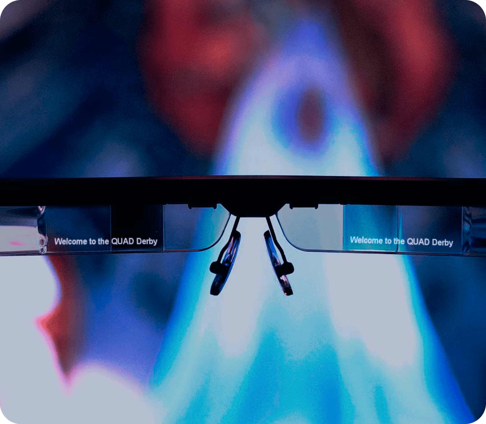 A close-up of smart glasses with clear lenses displaying the text Welcome to the QUAD Derby on both sides. The background is blurred with blue and red hues.