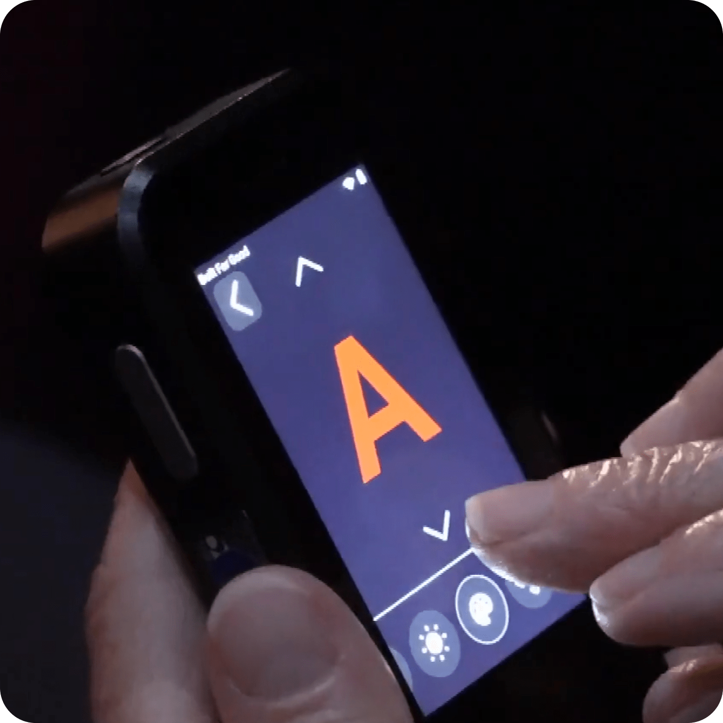A person uses a touchscreen device displaying a large orange letter A on a dark background, with navigation arrows and brightness control icons visible on the screen.