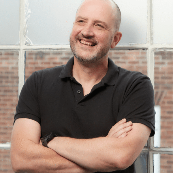 A smiling man with short hair and a beard, wearing a black polo shirt, stands with arms crossed in front of large frosted windows and a brick wall outside.