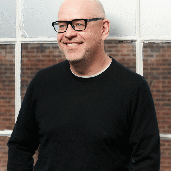 A smiling bald man with glasses, wearing a black sweater and standing in front of a window with a brick wall visible outside.