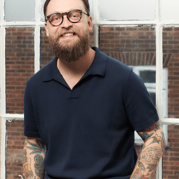 A smiling man with glasses, a beard, and tattoos on his arms sits in front of a window wearing a navy collared shirt and plaid pants.