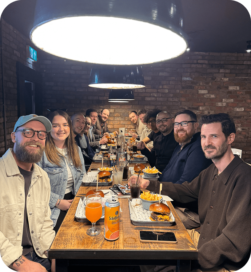 A group of people sit around a long wooden table in a restaurant, smiling at the camera. The table is filled with burgers, fries, and drinks. Two large circular lights hang above them, and a brick wall is in the background.