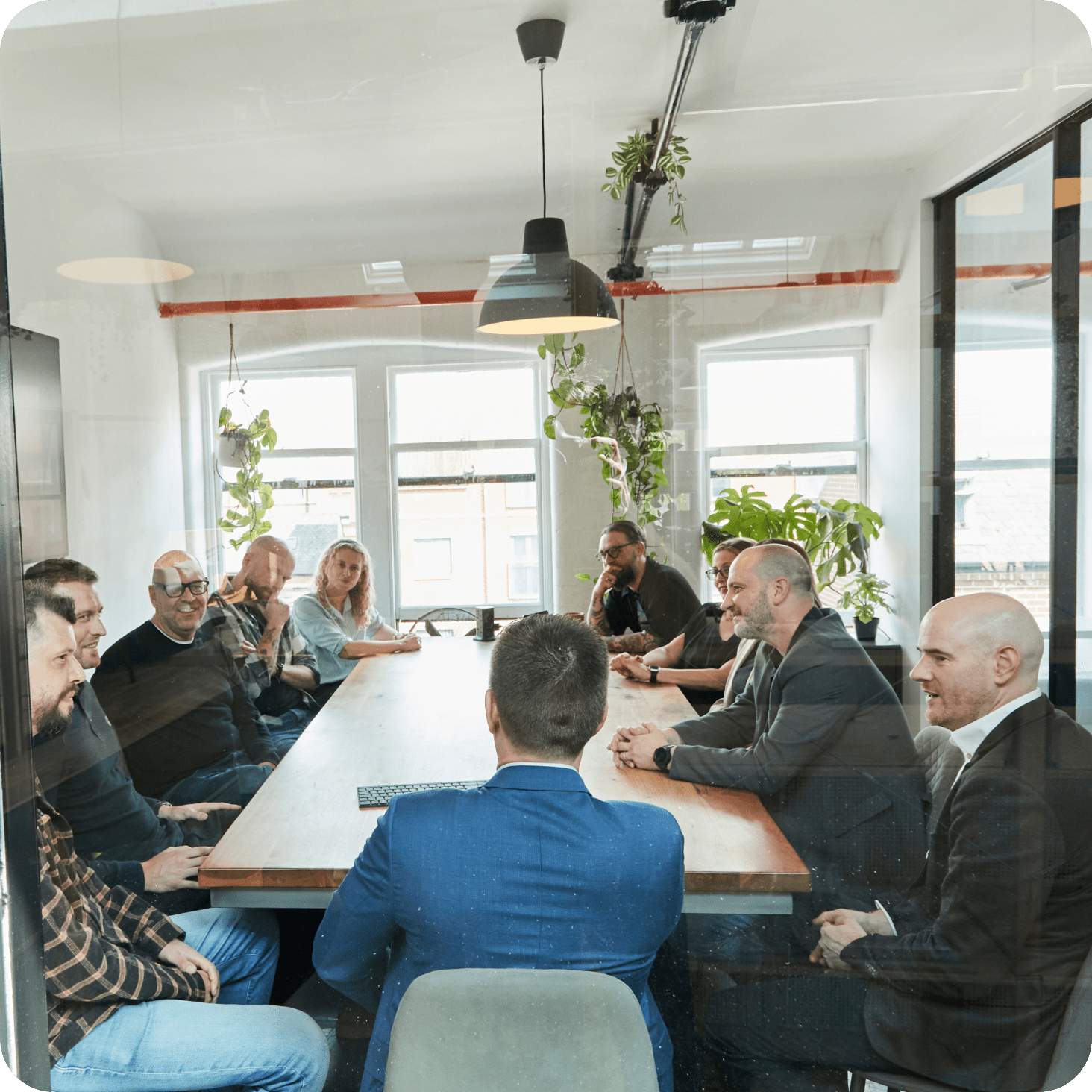A group of people sit around a large conference table in a modern office, engaged in a meeting. Some are smiling and conversing. The room is bright with large windows, plants, and minimalistic decor.