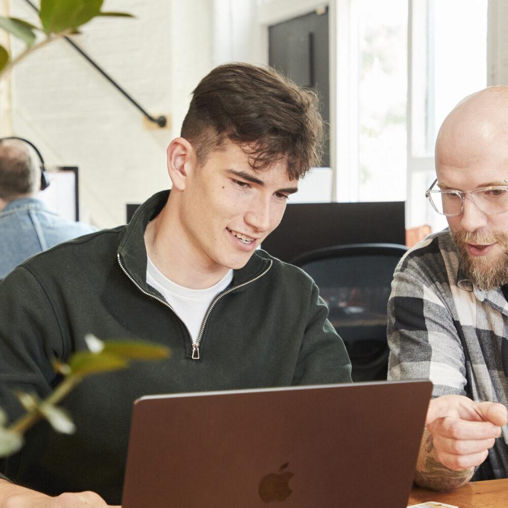 Two men sit at a desk in an office, looking at a laptop screen together. One man is pointing at the screen while the other smiles. Other people and plants are visible in the background.