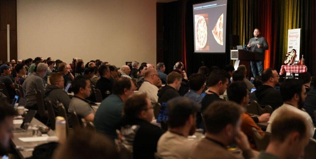 A speaker presents to a large seated audience at a conference. The screen behind shows an image of pizza slices. Attendees appear engaged, taking notes and using laptops. The room is well-lit with colorful curtains.