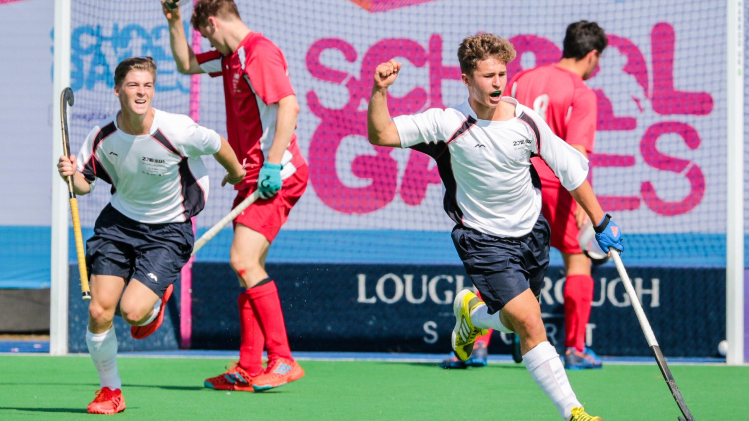 Two male field hockey players in white jerseys celebrate a goal, raising their arms and sticks, while players in red uniforms react near the goalpost on a bright green field. A “School Games” banner is visible in the background.