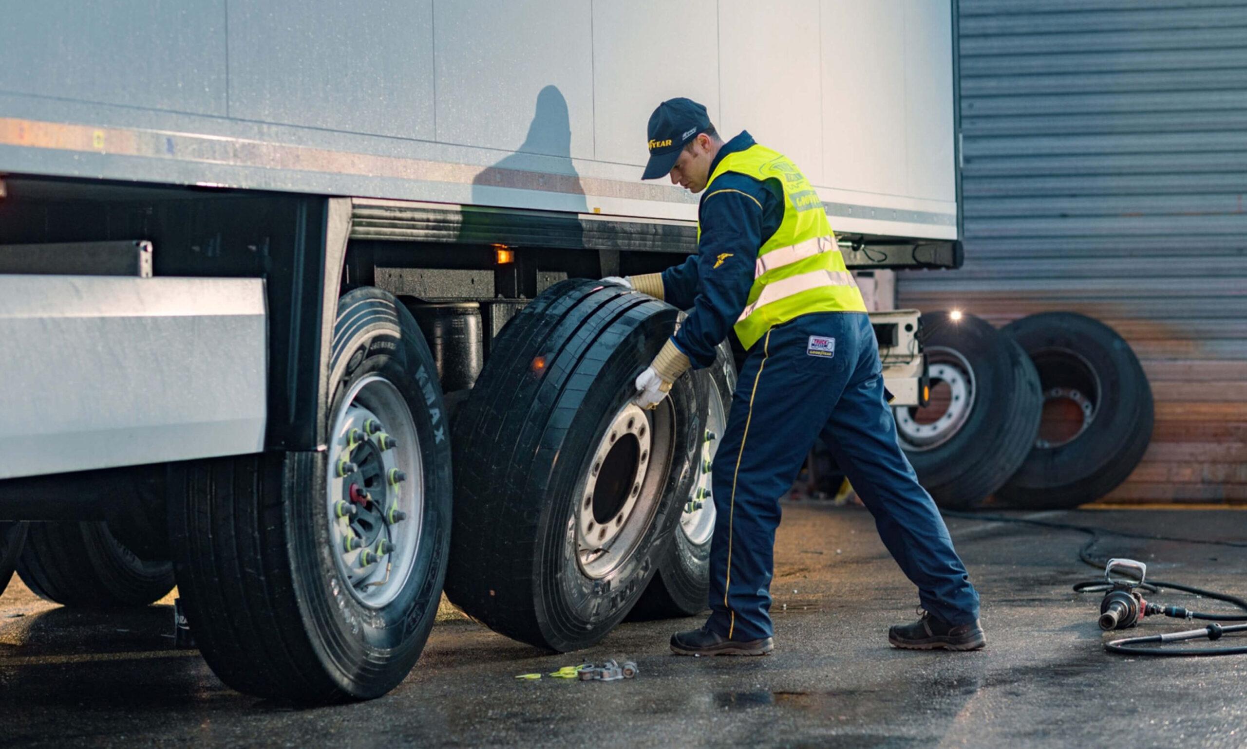 A worker in a yellow safety vest and cap is changing a large truck tire outside, standing next to the trucks rear wheels on a wet surface with tools and spare tires nearby.