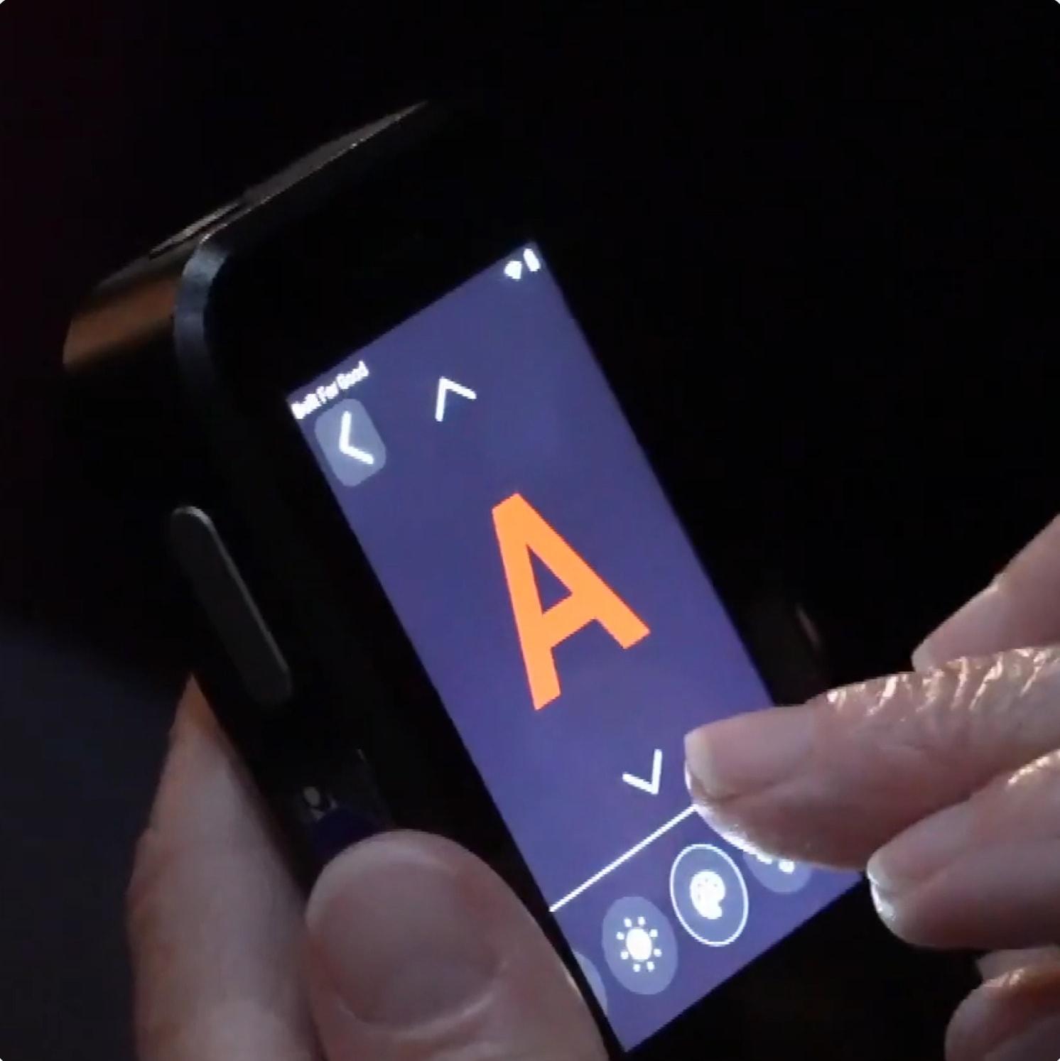 A person’s hand touches the screen of a handheld device displaying a large orange letter A with up and down arrows and brightness controls on the interface.