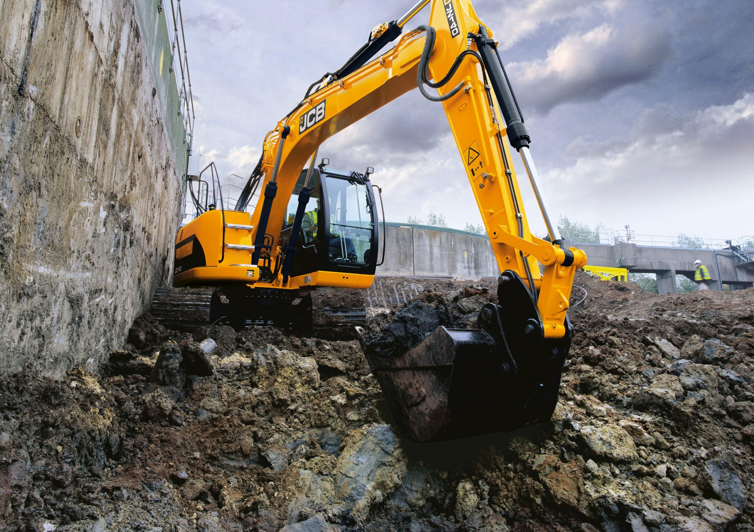 A yellow JCB excavator digs into rocky soil near a concrete wall at a construction site under a cloudy sky.